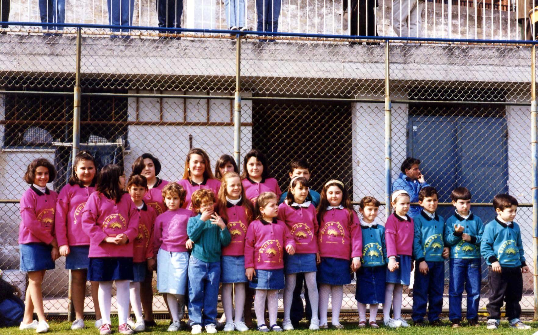 Bambini del coro in maglie rosa, riuniti all’aperto in un cortile.