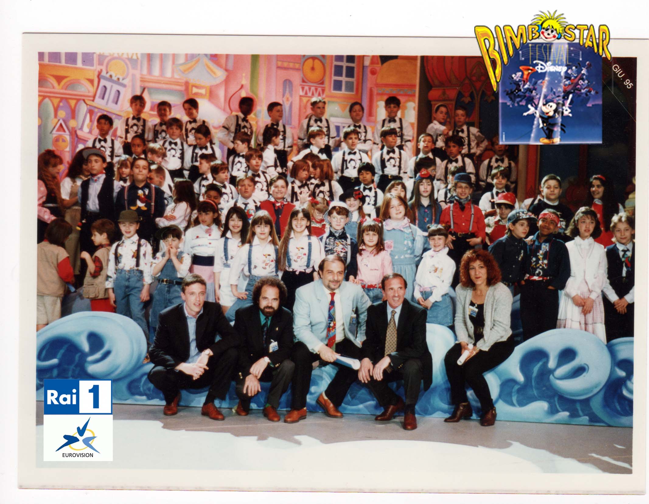 Foto di gruppo con numerosi bambini e cinque adulti in prima fila, davanti alla scenografia colorata del Festival Disney con onde azzurre.